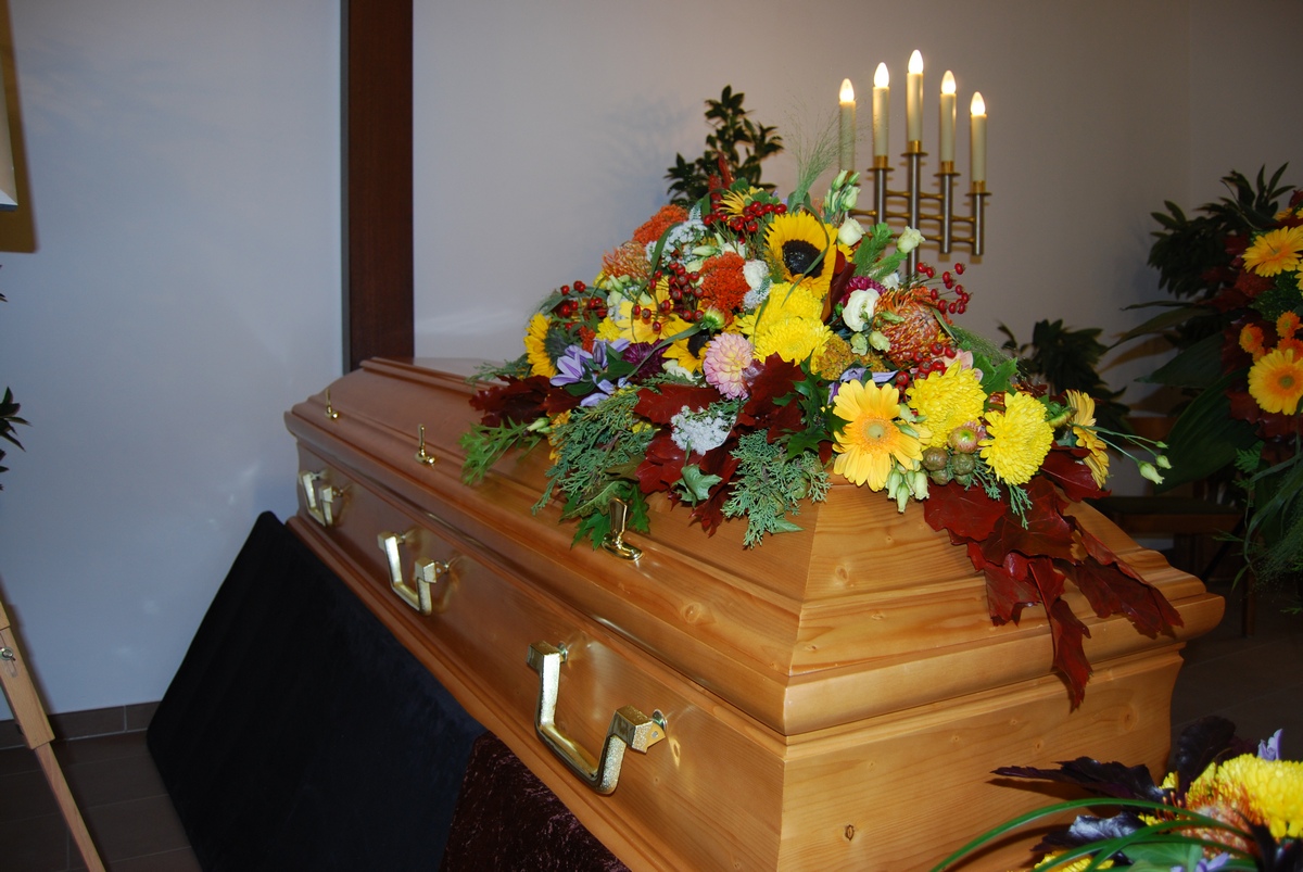 Sarg aus hellem Holz mit Sargbukett aus Sonnenblumen, Chrysanthemen und Herbstlaub
