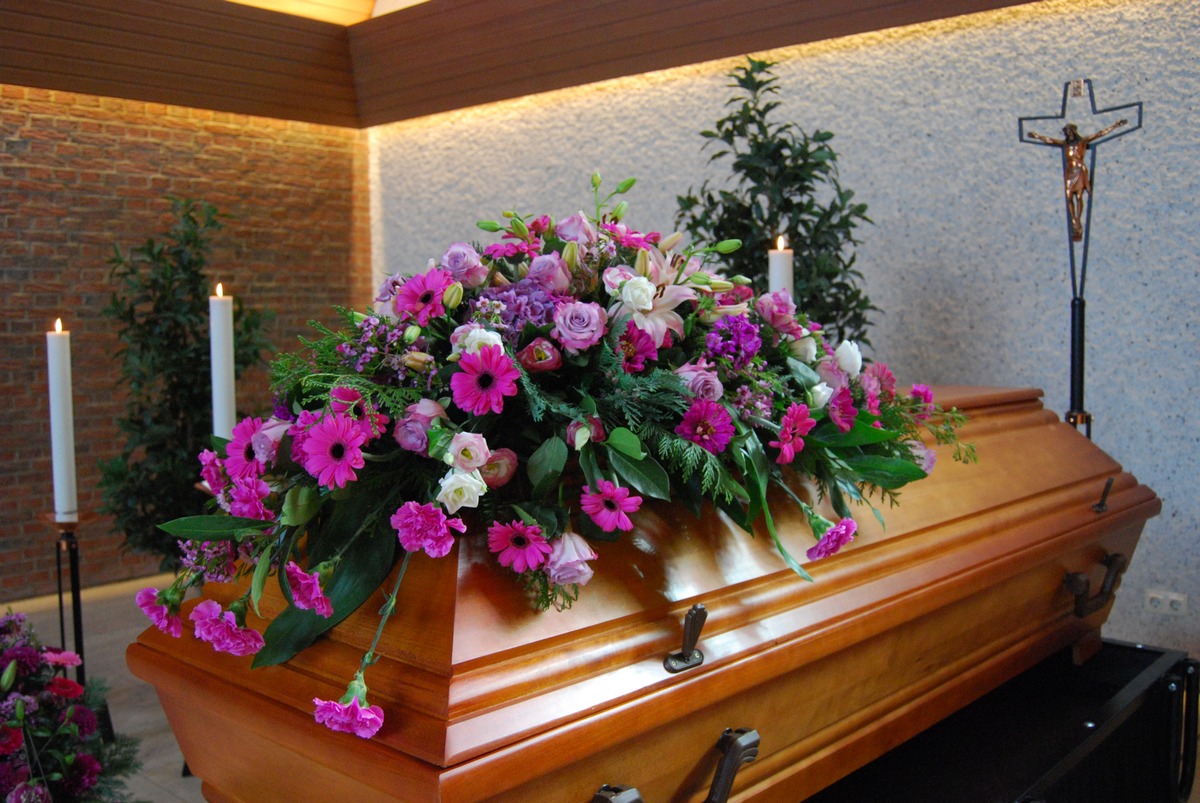Sarg im Aufbahrungsraum mit Sargbukett in Pink und Violett, Kerzen und Kreuz im Hintergrund