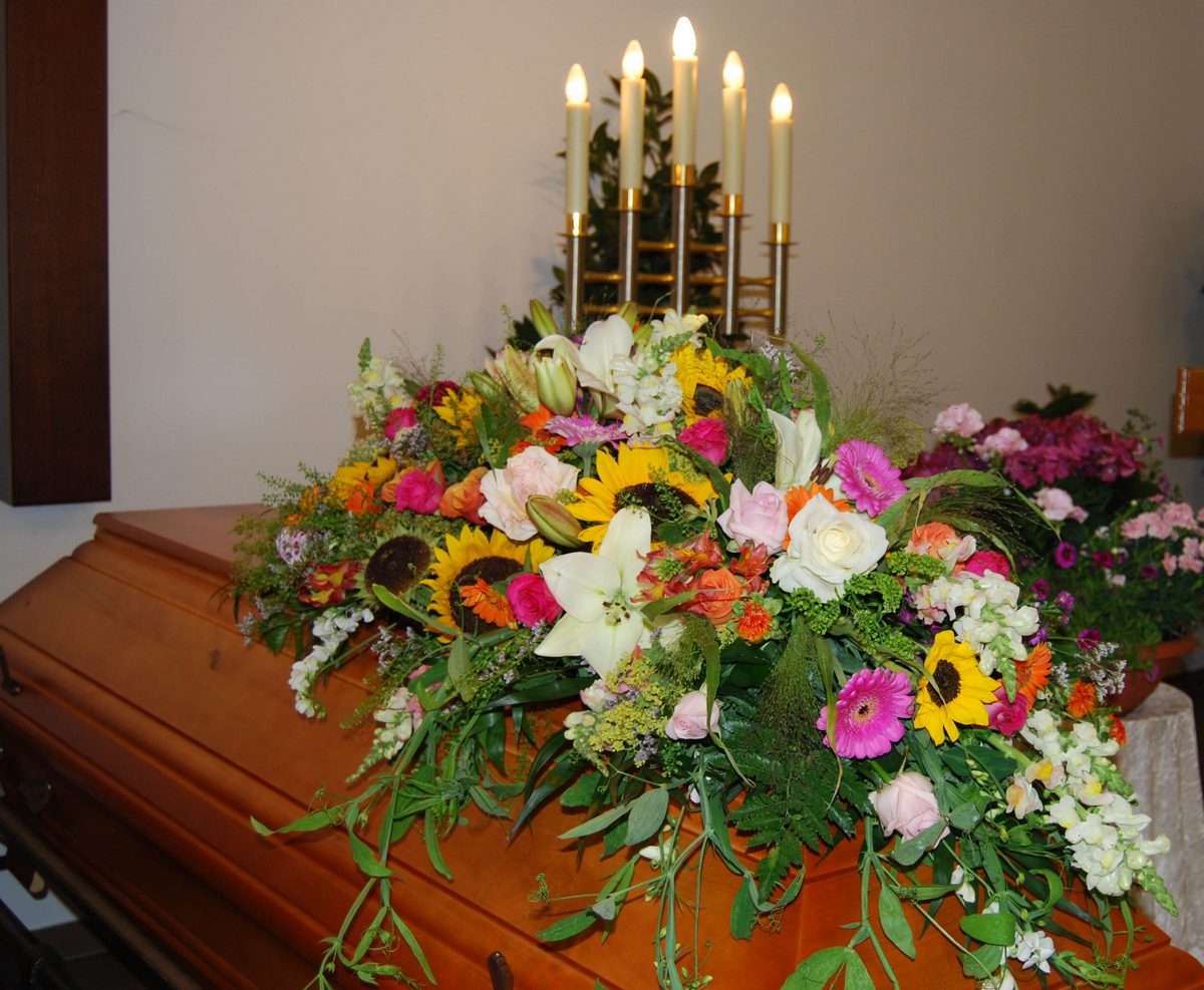 Sarg mit üppigem Sargbukett aus Sonnenblumen, Rosen und Gerbera vor einem Kerzenleuchter