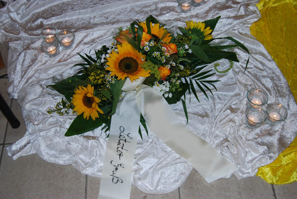 Liegendes Sonnenblumengesteck auf hellem Tuch mit Teelichtern und Trauerschleife