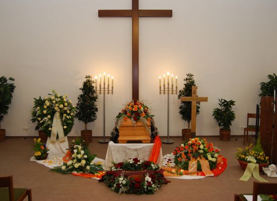 Trauerhalle mit Sarg vor einem Holzkreuz, Blumenschmuck in Weiß und Orange