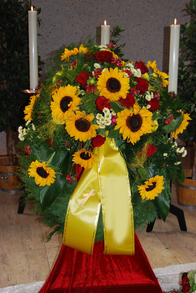 Trauerkranz mit Sonnenblumen und roten Rosen zwischen Kerzen, mit gelber Schleife