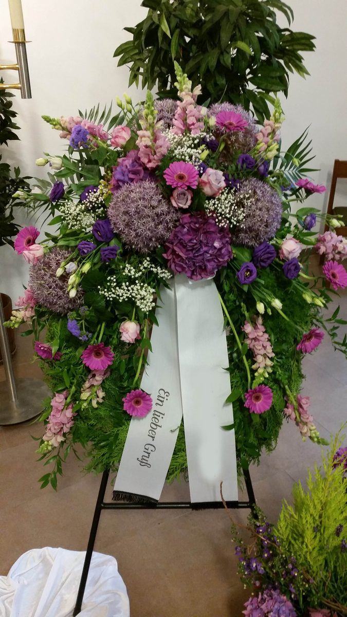 Trauerkranz in Violett und Rosa auf einem Kranzständer, mit heller Schleife