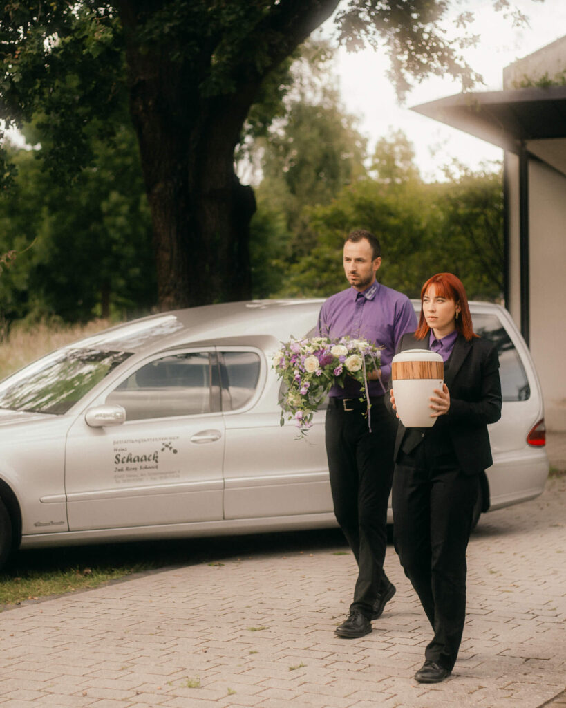 Zwei Mitarbeitende tragen Urne und Blumenschmuck zum Bestattungswagen von Bestattungen Heinz Schaack