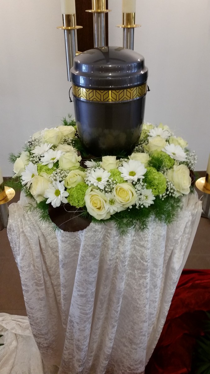 Dunkle Urne auf hellem Tuch mit einem Kranz aus weißen und grünen Blüten