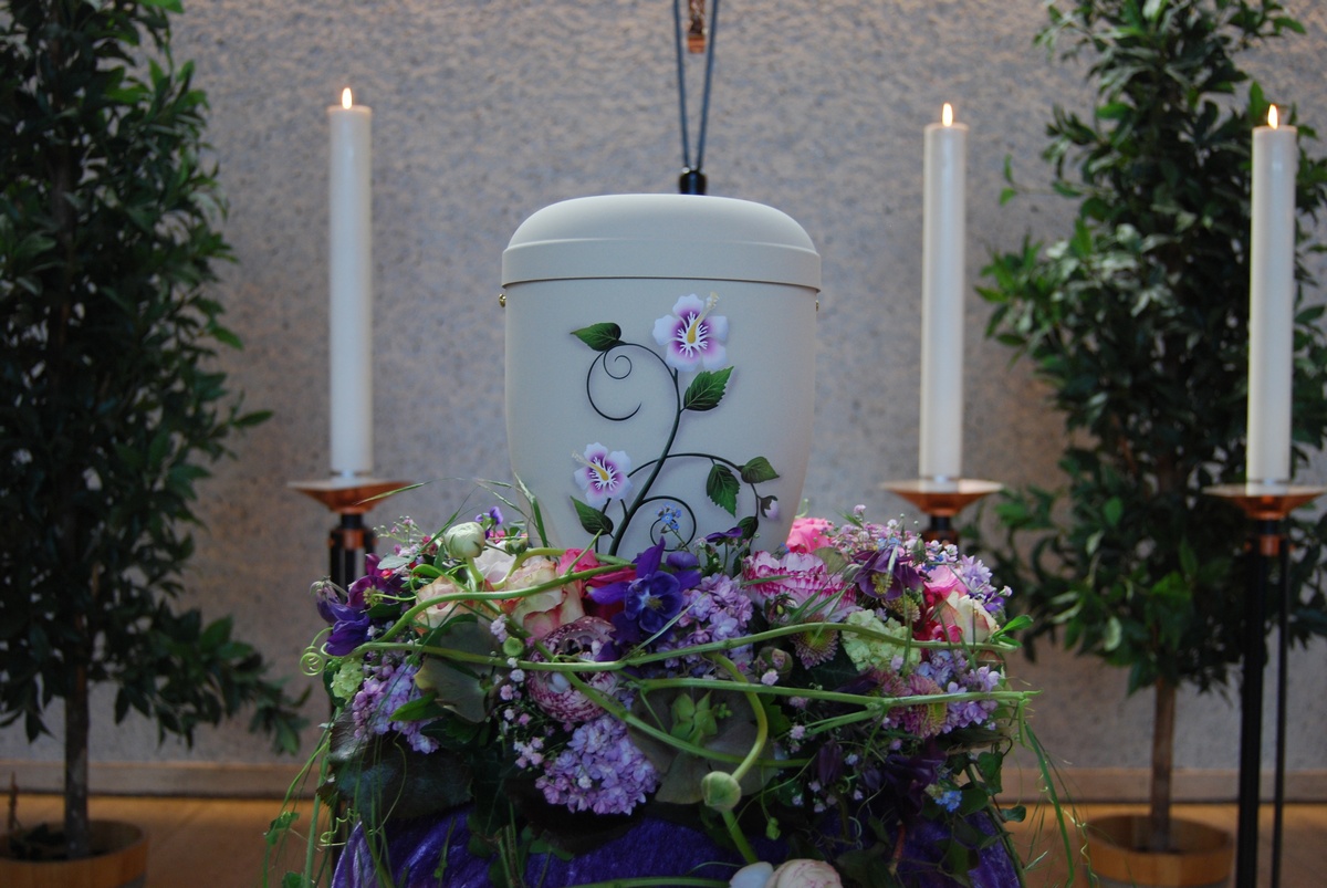 Weiße Urne mit Blumenmotiv, umgeben von Blütenschmuck in Violett und Rosa